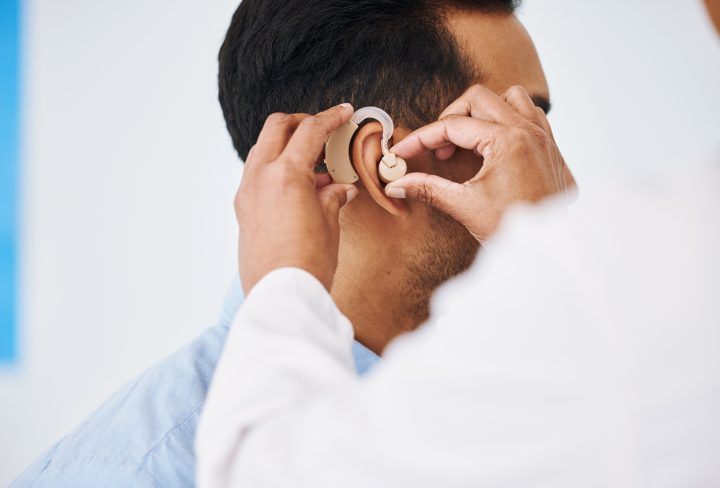 Doctor, man and hearing aid on ear for medical support, wellness and innovation of disability. Closeup, healthcare worker and deaf patient with audiology implant, service and help for sound waves.